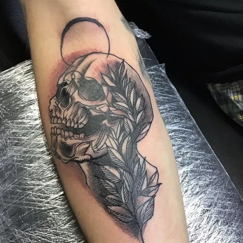 Tatuaje black and grey de una calavera entre hojas en el brazo.