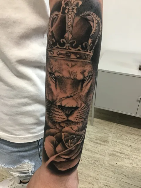 Tatuaje black and grey de un león con corona en el antebrazo.