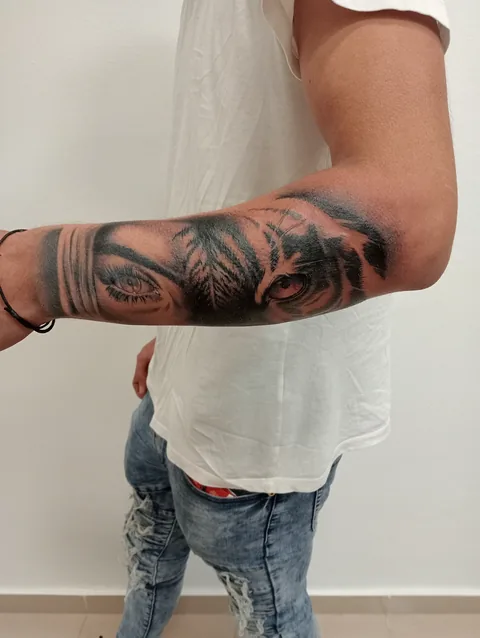 Tatuaje black and grey de unos ojos entre palmeras en el antebrazo.